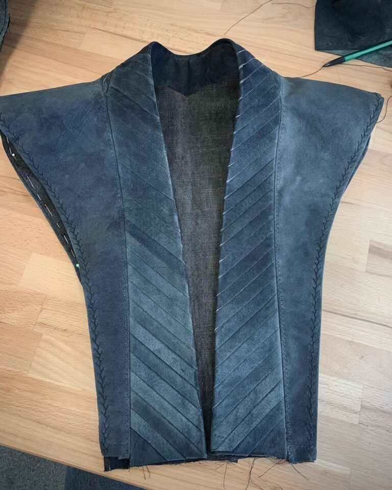 A suede jacket laid flat mid-construction, its front built from chevron-cut panels joined by hand-finished seams
